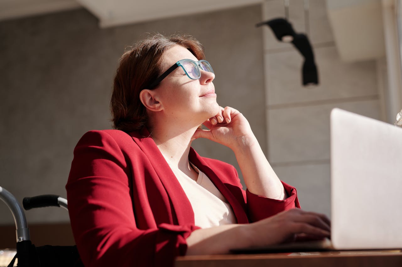 Woman in wheelchair wearing red suit and glasses, working on a laptop in a bright café.